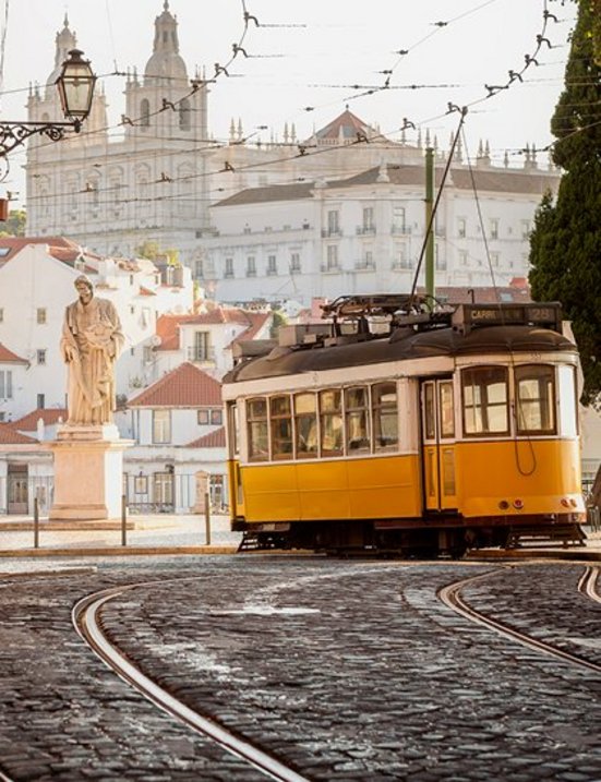 Eine gelb-weisse Strassenbahn fährt über eine Kopfsteinpflasterstrasse in Lissabon.