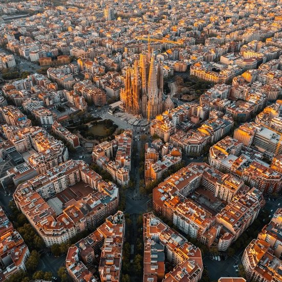 Luftaufnahme von Barcelonas Stadtbild bei Sonnenuntergang, mit warmen Farben und silhouettierten Gebäuden.