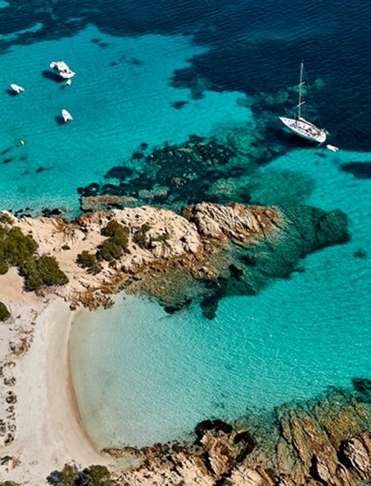 Strand in Sardinien mit Booten im Wasser und einer felsigen Küste im Hintergrund.
