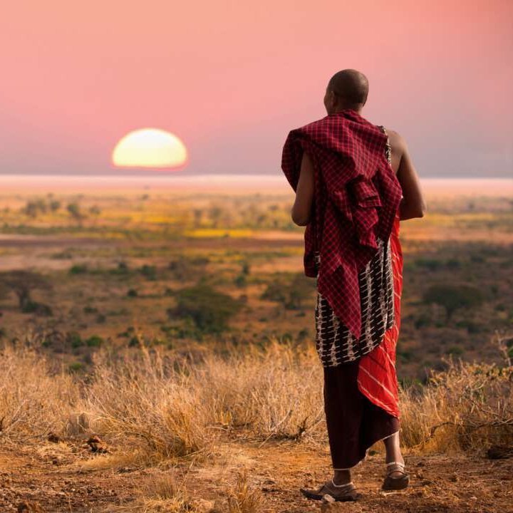 Ein Mann in einem roten Gewand steht bei Sonnenuntergang auf einer Ebene in Tansania.