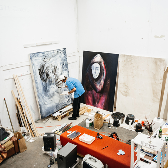 Ein Mann malt in einem Atelier in Berlin. Die Farben und Pinsel liegen um ihn herum verstreut.