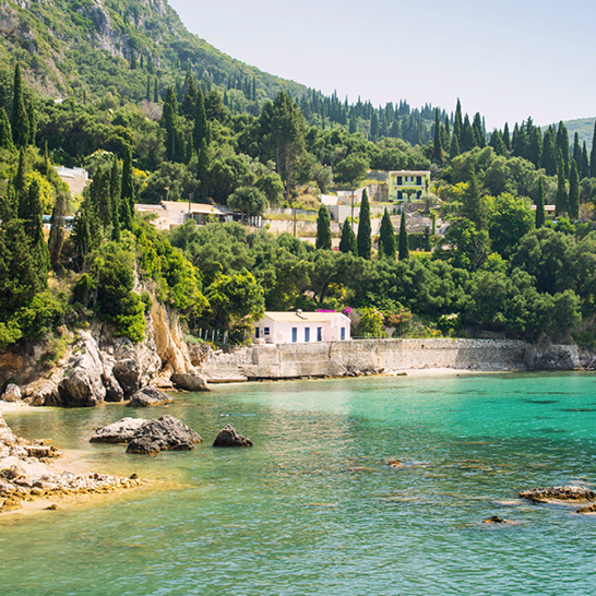Kleines weisses Haus am Ufer eines Gewässers auf Korfu, umgeben von Natur und sanften Wellen.