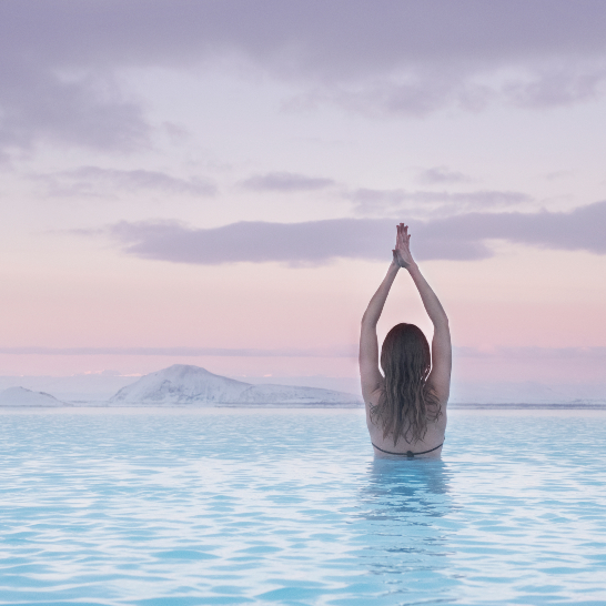 Frau im blauen Lagunenwasser bei Sonnenuntergang, umgeben von sanften Wellen und warmen Farben des Himmels.