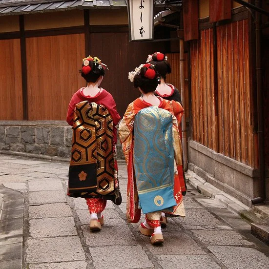 In Japan spazieren drei Geishas durch eine enge Gasse, ihre Kimonos strahlen in lebhaften Farben.