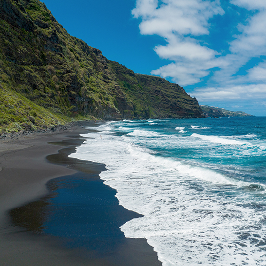  Plage de sable noir aux Iles Canaries, avec une montagne majestueuse en arrière-plan sous un ciel ensoleillé.