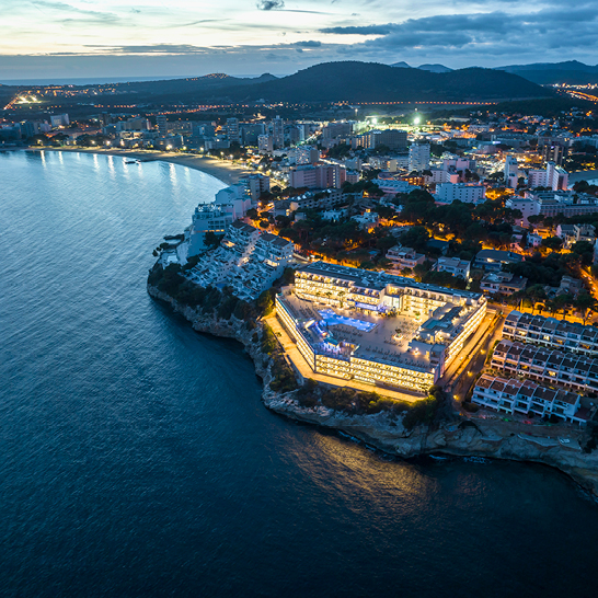 Luftaufnahme eines Hotels und Strandes bei Dämmerung auf Mallorca, mit sanften Wellen und einem farbenfrohen Himmel.