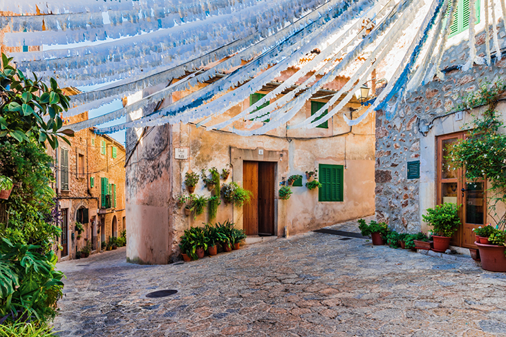 Eine Strasse auf Mallorca, gesäumt von vielen Pflanzen und geschmückt mit blauen und weißen Fahnen.
