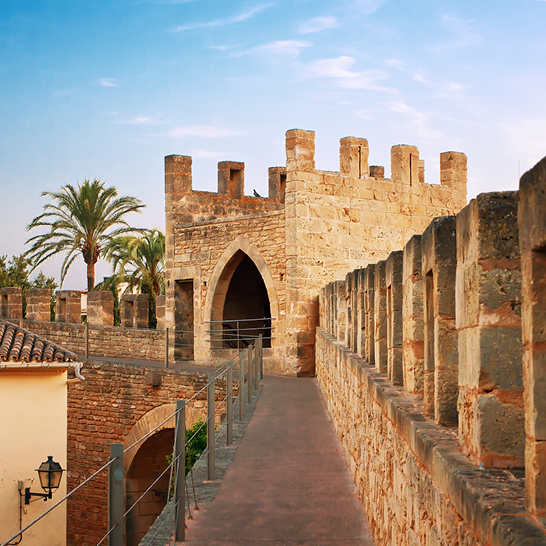 Eine Burg mit einem Uhrturm und einer Palme in Alcudia, umgeben von blauem Himmel und grüner Landschaft.