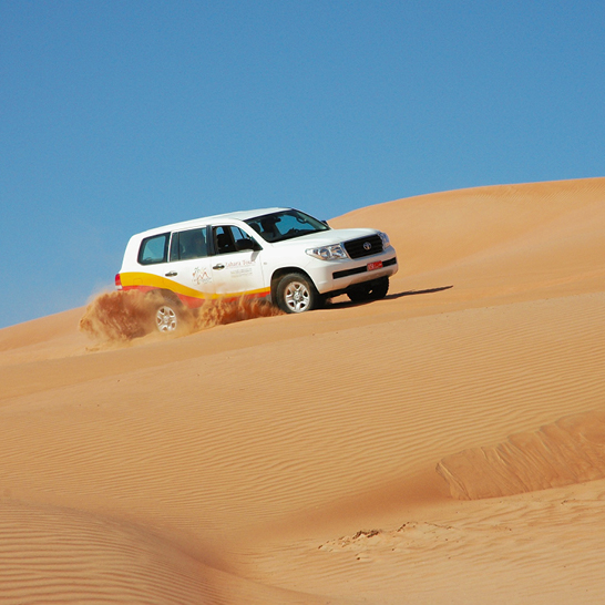 Ein weisser SUV fährt durch die sandige Wüste im Oman, umgeben von sanften Dünen und strahlend blauem Himmel.