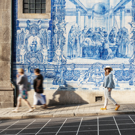 Eine Frau spaziert auf der Straße, während die Sonne scheint und Passanten vorbeigehen.