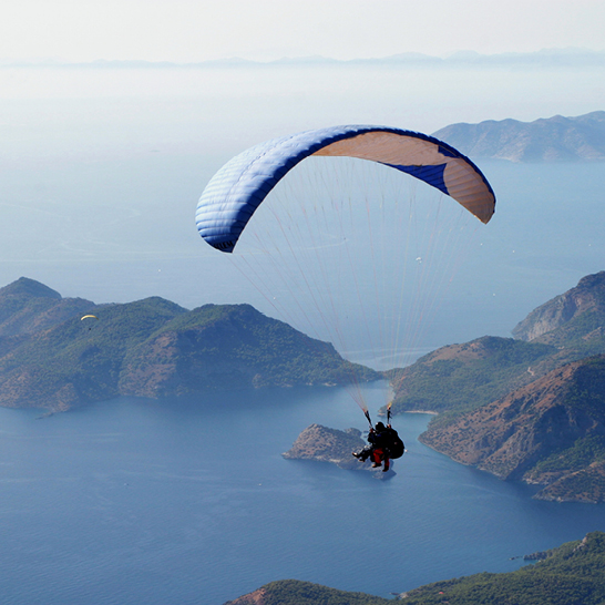 Eine Person fliegt mit einem Gleitschirm über ein Gewässer in der Türkei, umgeben von atemberaubender Landschaft.