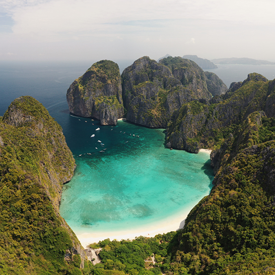 Vue d'une plage en Thaïlande avec une chaîne de montagnes en arrière-plan, ciel bleu et vagues douces.