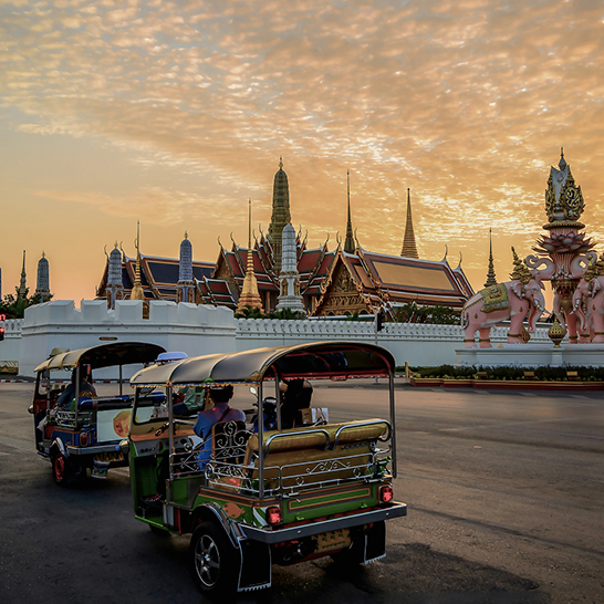 Deux tuk-tuks garés sur une rue animée de Bangkok, entourés de bâtiments colorés et de passants.